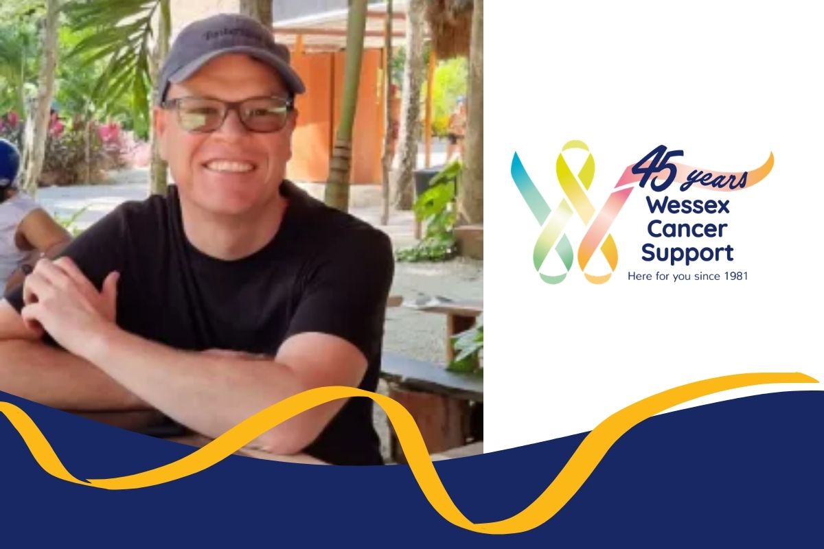 Photo shows Deon wearing a black t-shirt and a cap and smiling to camera, with his arms folded in front of him.